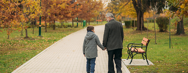 Älterer Mann spaziert im Herbst mit seinem Enkel in einem Park auf einem Weg mit Bänken.
