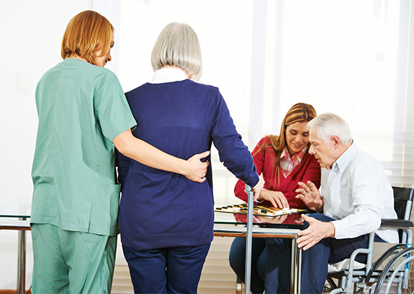Pflegerin begleitet eine Bewohnerin in einem Pflegeheim und älterer Mann im Rollstuhl bei einer Aktivität mit einer Angehörigen.